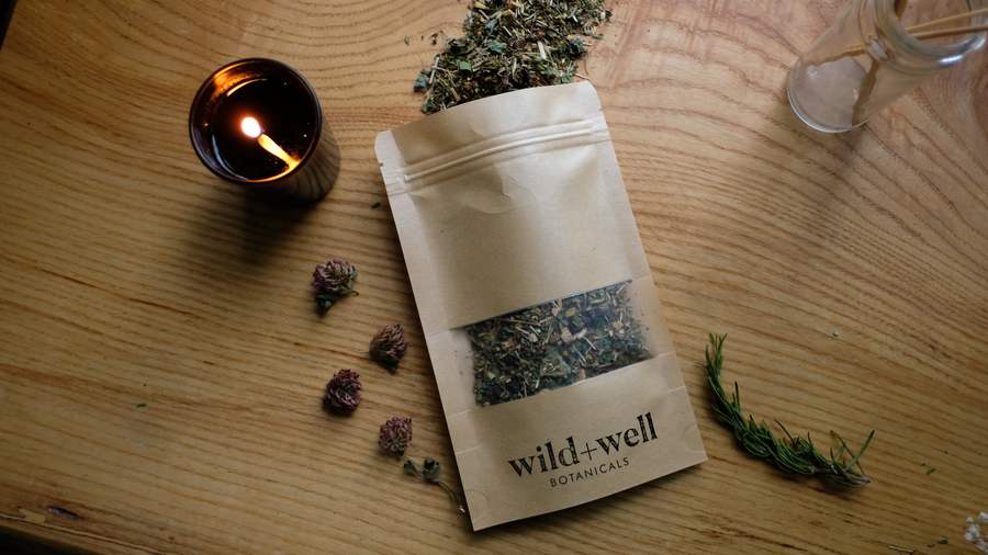 Tea pouch with loose tea spilling out, beside a lit candle, clover and sprigs of rosemary