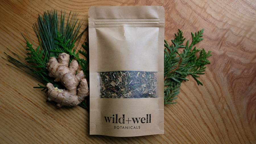 Kraft paper tea pouch beside fresh ginger root and cedar sprigs on an oak table